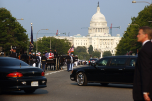 Trauerzug mit Menschen in Mützen und Fahnen, Fahrzeugen, Laternenpfählen, Bäumen und Gebäuden mit Fenstern vor dem Kapitolsgebäude unter einem klaren Himmel.