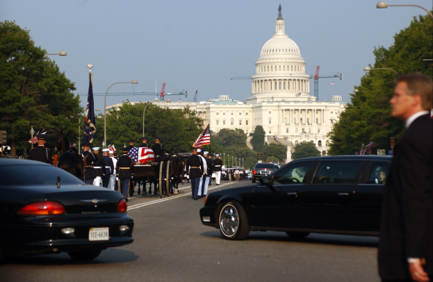 Trauerzug mit Menschen in Mützen und Fahnen, Fahrzeugen, Laternenpfählen, Bäumen und Gebäuden mit Fenstern vor dem Kapitolsgebäude unter einem klaren Himmel.
