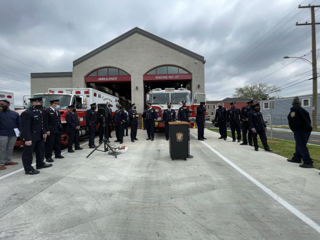 Gruppe von Menschen in Uniformen neben einem Feuerwehrauto auf einem Podium während einer Feuerwache-Eröffnung, mit Gebäuden, Bäumen und Strommasten im Hintergrund.