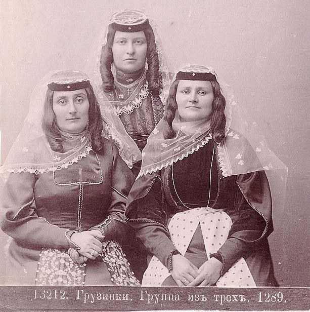 Drei Frauen in traditionellen russischen Hochzeitskleidern mit farbenfroher Stickerei und Perlen, die vor einer weißen Wand in einem Schwarz-Weiß-Foto posieren.