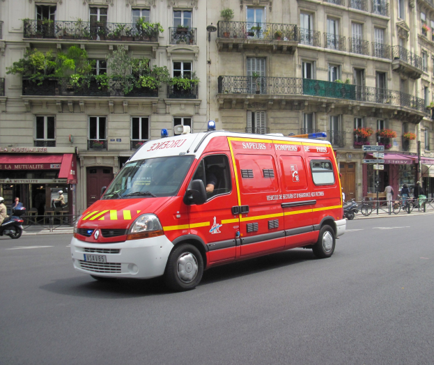Ambulanz auf dem Seitenstreifen vor einem Gebäude mit Fenstern, Geländern, Pflanzen und anderen Gegenständen geparkt, mit ein paar Menschen und Fahrrädern im Hintergrund.