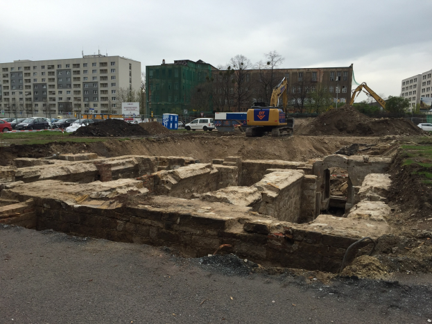 Großer Schutthaufen auf einer Baustelle umgeben von Baggern, Fahrzeugen, Gras, Bäumen und Gebäuden mit Fenstern in einem bewölkten Himmel über einer Straße.