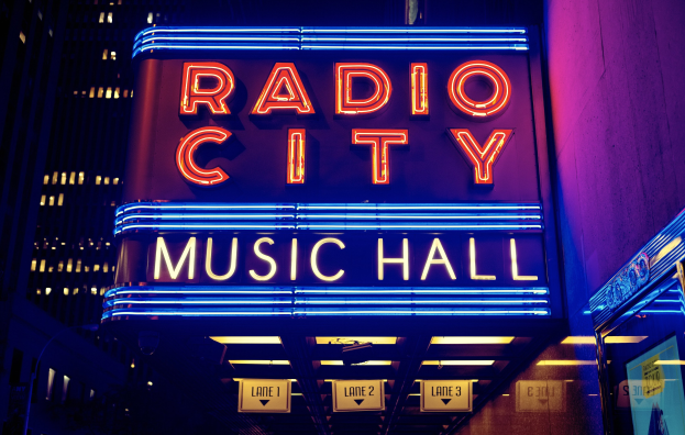 Hell erleuchtetes Radio City Music Hall in New York City, flankiert von hohen Gebäuden, mit einem "Radio City"-Schild und zusätzlichen Schildern auf der rechten Seite.
