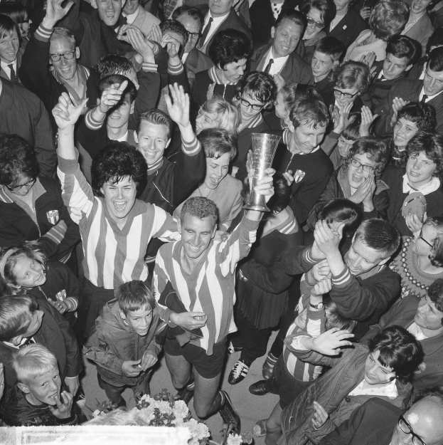 Schwarzes Bild einer Gruppe von Menschen, die mit einem Pokal feiern, Blumen unten und einer Menge im Hintergrund.