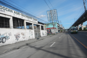 Eine Straße in einer Stadt mit Fahrzeugen, eine Brücke auf der rechten Seite, Gebäude, Strommäste mit Käblein und einen bewölkten Himmel, mit einem graffiti-beschmierten Gebäude an der Straßenseite.