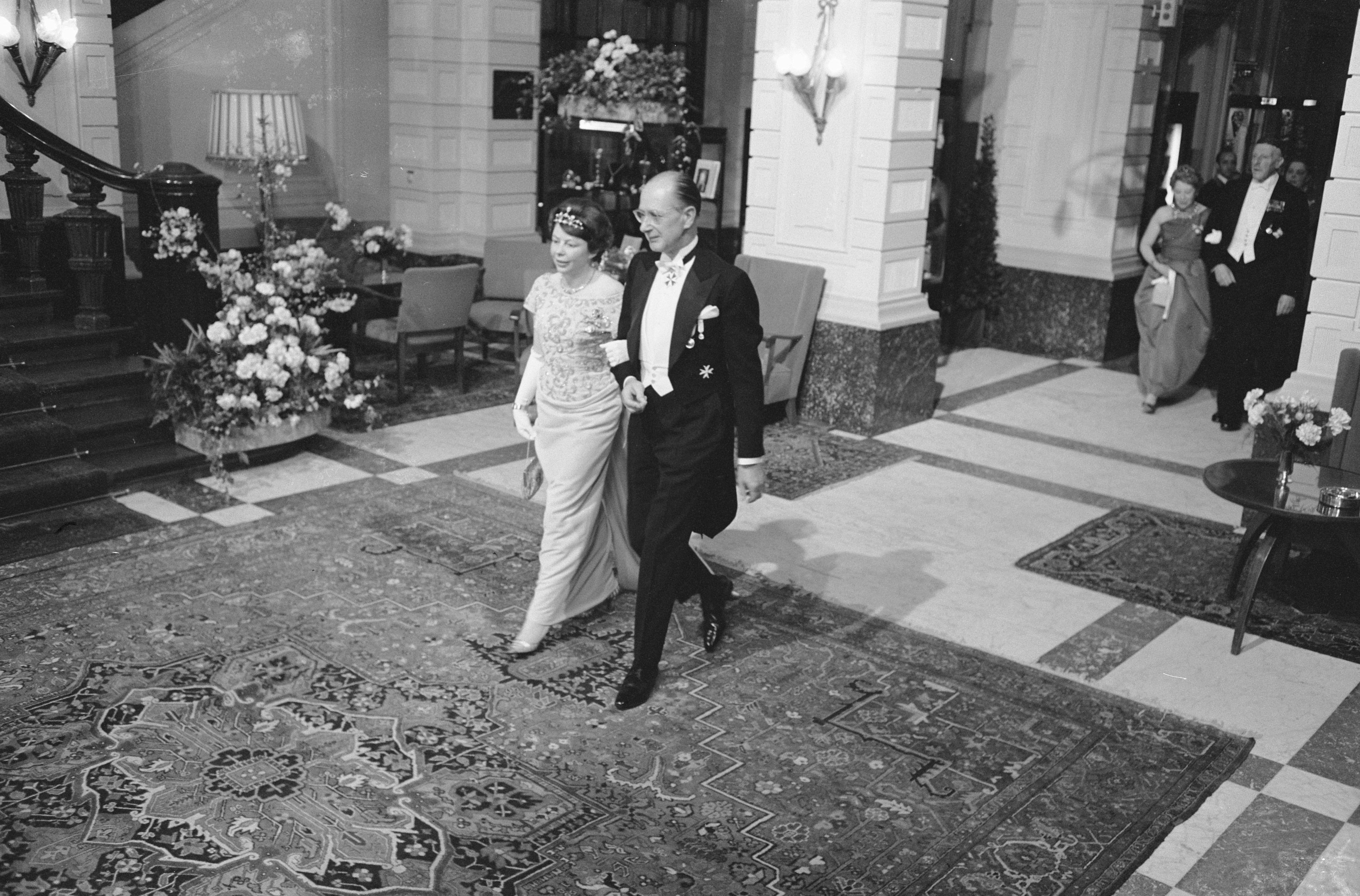 Schwarz-weiß-Foto einer Braut und eines Bräutigams, die durch die Lobby eines Hotels gehen, umgeben von Gästen, mit Blumenschmuck, Möbeln und architektonischen Details