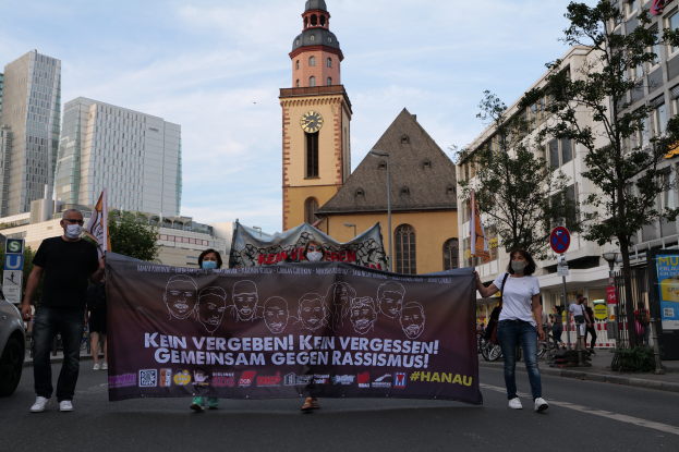 Eine Gruppe maskierter Menschen marschiert eine Straße entlang und hält ein Transparent in der Hand, mit einem geparkten Auto auf der linken Seite, Gebäuden, Bäumen, Schildern, Pfählen und einem Kirchturm im Hintergrund unter einem klaren blauen Himmel.