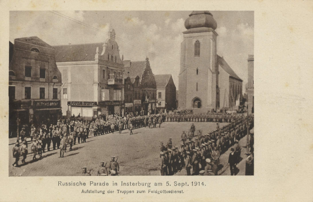 Schwarzes und weißes Foto einer Parade in Insterburg im Jahr 1914 mit marschierenden und stehenden Menschen, Gebäuden im Hintergrund und Wolken am Himmel.