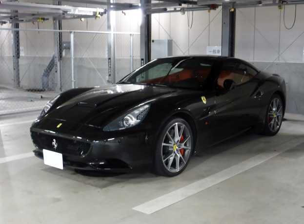 Schwarzer Ferrari California in einer Parkgarage mit einer Wand im Hintergrund, Maschendraht auf der linken Seite und Deckenlampen.