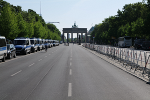 Lange Reihe von Polizeiwagen entlang einer Straße vor dem Brandenburger Tor geparkt, mit Fahrradfahrern und Füüspersonen, Absperrungen, Bäumen und einem Bogen mit Statuen im Hintergrund.