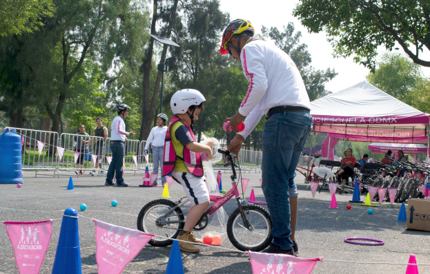 Ein Mann hilft einem jungen Mödchen in einem rosafarbenen und weißen Kleid und einem weißen Helm beim Radfahren in einem Park, mit anderen und Fahrrädern im Hintergrund.