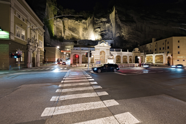 Eine nächtliche Stadtstraße mit fahrenden Autos, Straßenlaternen, Ampeln, Gebäuden, Laternenmasten, einem Hügel im Hintergrund und einer Zebrastreifenfurt am Boden.
