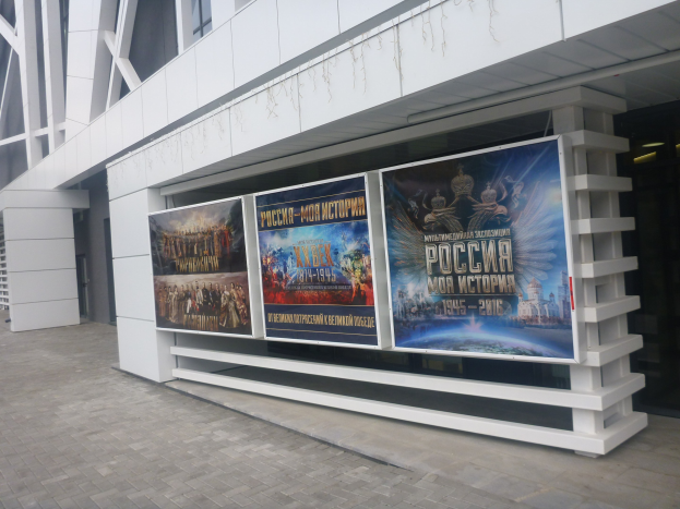 Großes Plakat an einem Gebäude, das die Eröffnungszeremonie der Olympischen Spiele in Moskau bewirbt, mit beleuchteter Gebäudeseite und sichtbarem Boden.