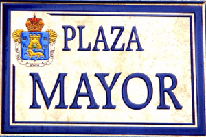 Rechtwinkliges weißes Schild mit schwarzer Schrift "Plaza Mayor" in fetter Schrift, blauem Rand und einem gekrönten Wappen-Logo an einer Gebäudefassade.