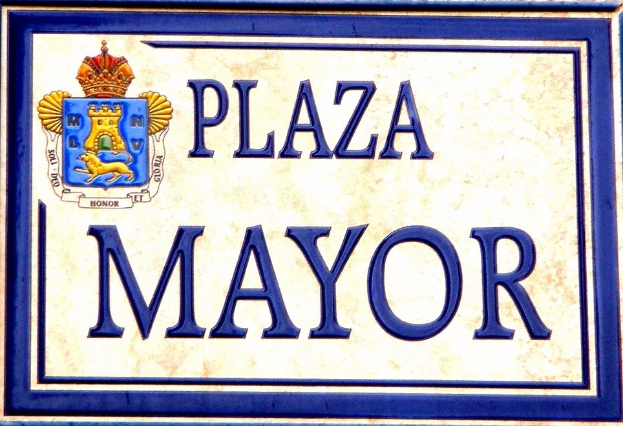 Rechtwinkliges weißes Schild mit schwarzer Schrift "Plaza Mayor" in fetter Schrift, blauem Rand und einem gekrönten Wappen-Logo an einer Gebäudefassade.