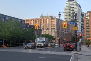 Eine städtische Straße mit starkem Verkehr, einschließlich eines Lastwagens, eines Autos und eines SUV, gesäumt von Laternenmasten, Verkehrsampeln, Texttafeln, Bäumen und Gebäuden unter einem Himmel.