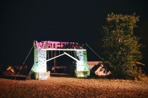 Bühne für ein nächtliches Musikfestival mit einem Tor mit Text, einem Baum auf der rechten Seite, Zelten im Hintergrund und einem dunklen Himmel.