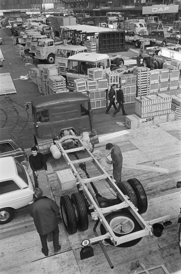 Schwarze und weiße Fotografie von Männern, die an einem Truck in einer Fabrik arbeiten, mit Fahrzeugen, Menschen und Kisten im Hintergrund.