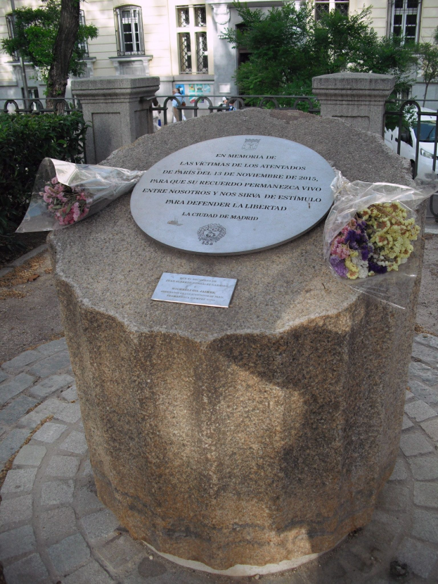 Steindenkmal mit einer Plakette, umgeben von Blumensträußen, vor dem Hintergrund von Gebäuden, Bäumen, Geländern, Fahrzeugen und Menschen.