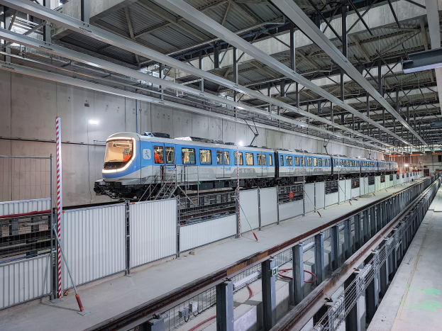 Blauer und weißer Zug fährt durch eine große Halle mit einer Plattform auf der rechten Seite und einer Wand im Hintergrund, unter einer Decke mit Stangen und Lampen.