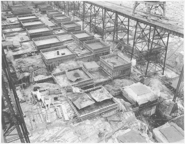 Ein Schwarz-Weiß-Foto einer Baustelle mit einer Brücke im Hintergrund, das verstreute Gebäude, Metallstangen und andere Gegenstände zeigt, mit Text am unteren Bildrand.