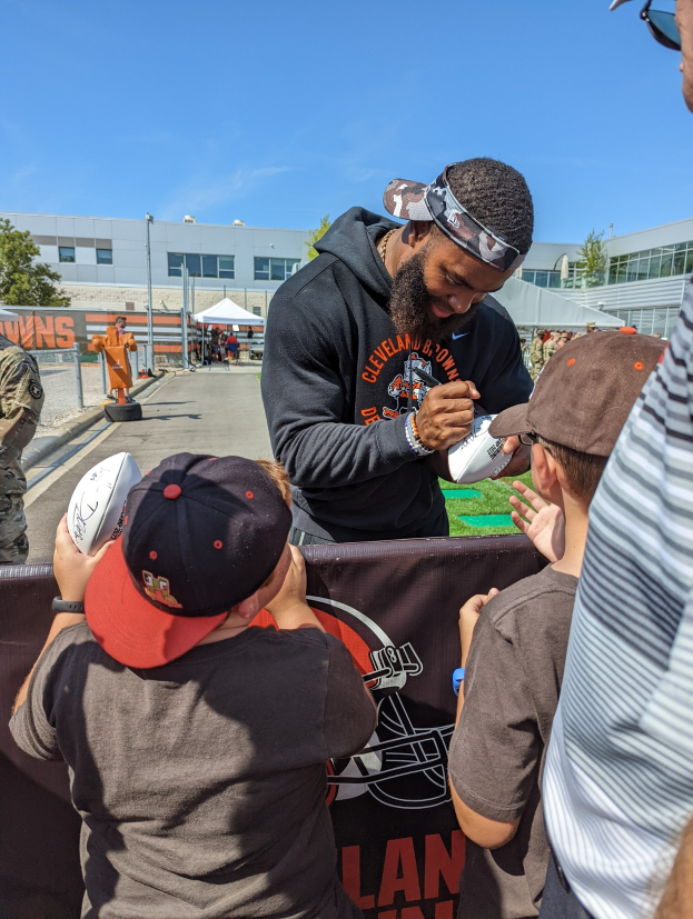 Cleveland Browns Wide Receiver Odell Beckham Jr. signiert Autogramme für Fans auf dem Trainingsgelände, umgeben von Menschen mit Kappen, einem Banner und Gebäuden im Hintergrund unter einem bewölkten Himmel.