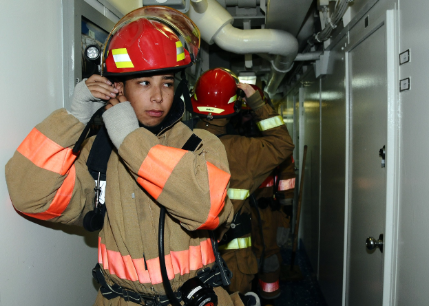 Feuerwehrleute in Uniform in einem Trainingsraum mit Rohren und Equipment im Hintergrund.