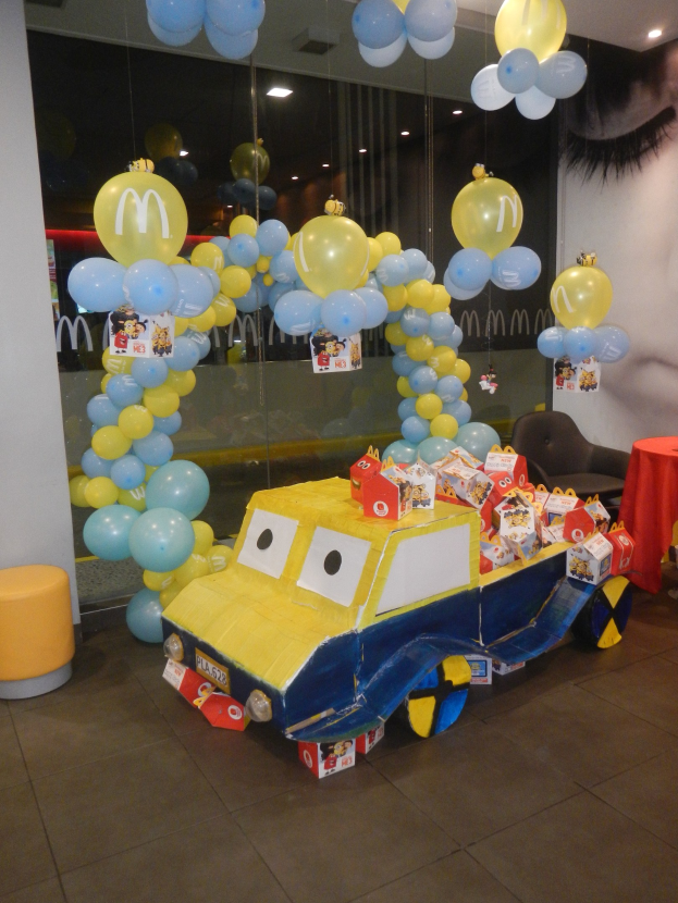 Eine McDonald's-Geburtstagsfeier mit einem gelben und blauen Lastwagen, der mit Ballons und anderen Dekorationen geschmückt ist, einem roten Tisch mit Gegenständen, Hockern, einer Glaswand und Deckenlampen, die eine festliche Atmosphäre schaffen.