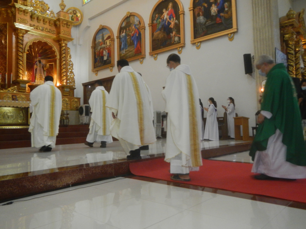 Eine Gruppe von Priestern geht über einen roten Teppich in einer Kirche, während Menschen auf dem Boden vor und rechts von ihnen stehen und Fotorahmen an der Hintergrundwand hängen.