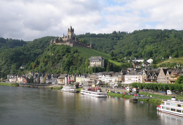 Ein malerischer Blick auf den Rhein in Deutschland, mit einer Burg auf einem Hügel, Booten auf dem Fluss, Fahrzeugen auf einer nähergelegenen Straße und einem bewölkten Himmel.