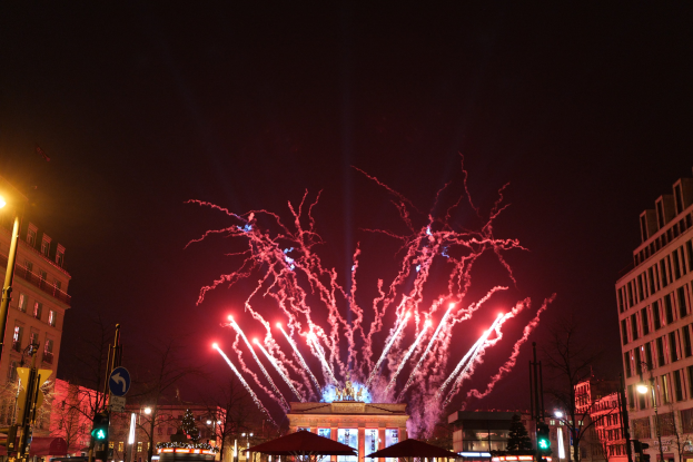Eine belebte Stadtstraße in Berlin am Silvesterabend, geschmückt mit Lichtern, Feuerwerk und umgeben von Gebäuden, Fahrzeugen und Passanten.
