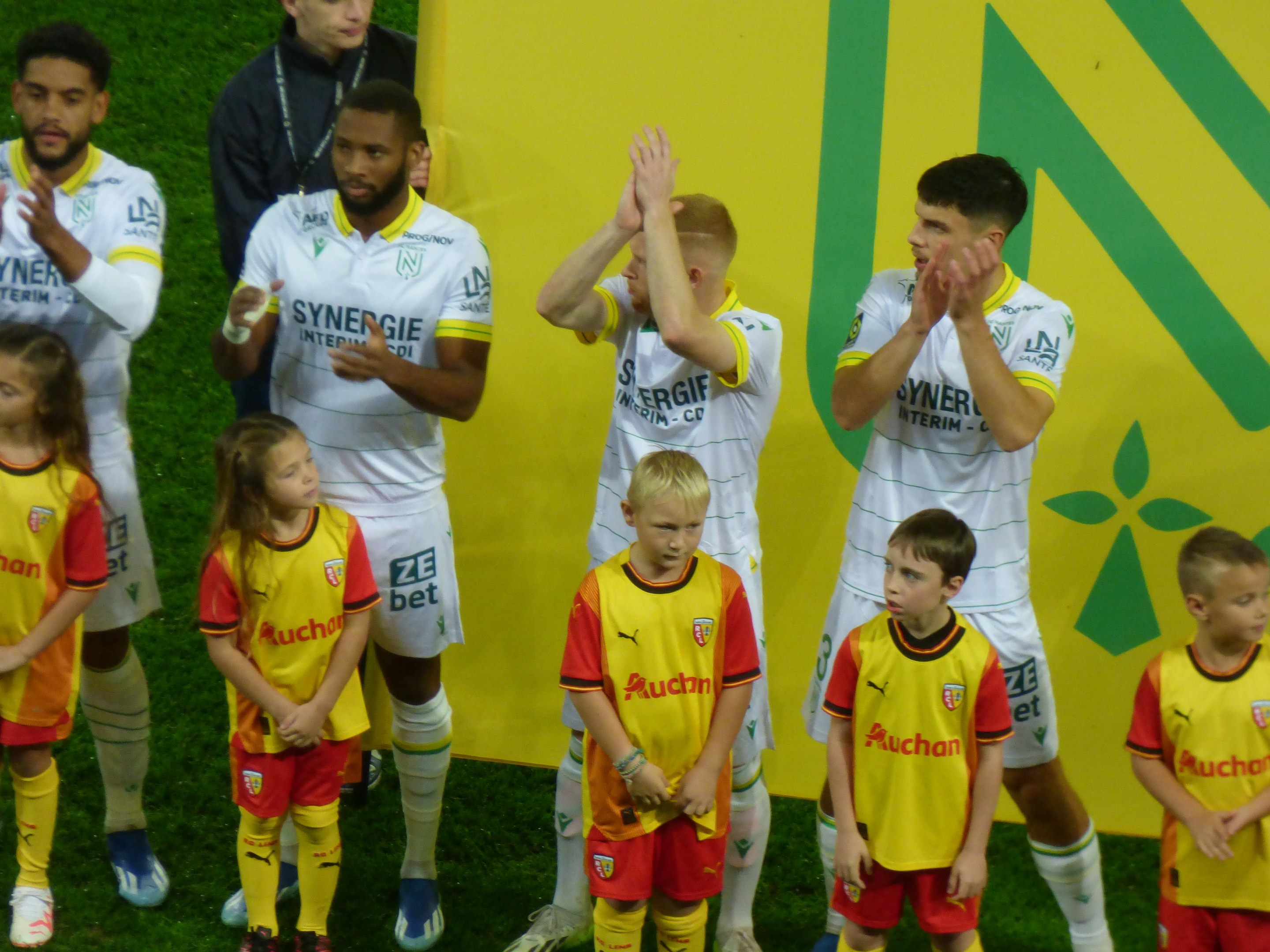 Eine Gruppe von Fußballspielern in Uniformen, die auf einem Feld klatschen, mit einem "Ligue 1"-Schild im Hintergrund, feiern einen Sieg.