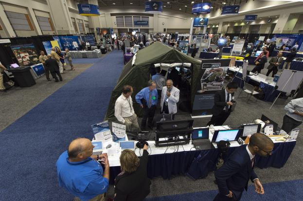 Große Gruppe von Menschen auf einer Karriere-Messe um Tische mit Laptops, Monitoren und Papieren versammelt, mit einem Zelt, Tafeln, Deckenleuchten und Fenstern im Hintergrund.