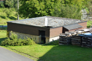 Kleines Holzhaus neben einer Straße, umgeben von Gras, Pflanzen, Bäumen und einem Pfahl, mit gestapelten Holzscheiten auf der rechten Seite.