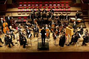 Großes Orchester auf der Bühne mit dirigierendem Musiker, Musikständer und Hintergrundtreppe und -stühlen.