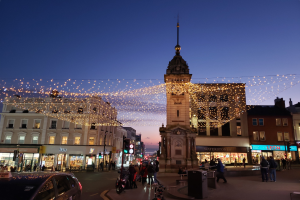 Eine belebte Stadtstraße mit Fußgängern, Fahrzeugen, Fahrrädern, Mülltonnen, Gebäuden, Laternen, Verkehrsampeln und einem Uhrenturm im Hintergrund, geschmückt mit Weihnachtslichtern.