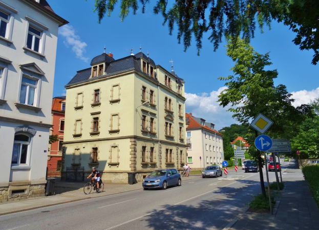 Eine Stadtstraße mit Autos und Fahrradfahrern, hohe Gebäude mit Fenstern, Schilder an Pfählen, Bäume und ein bewölkter Himmel.