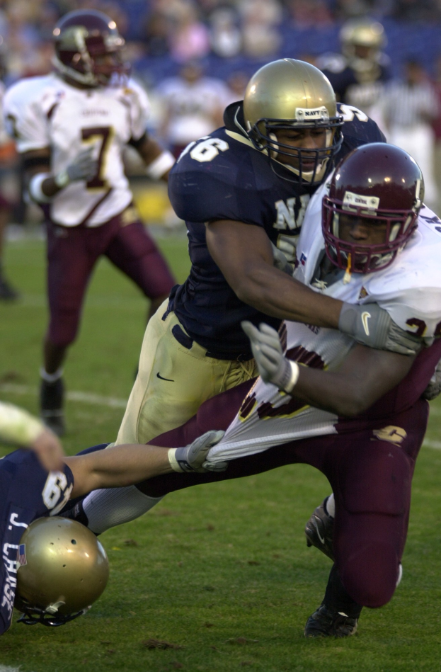 Zwei Fütballspieler in Helmen und Uniformen stoßen auf dem Feld zusammen, während andere zuschauen.