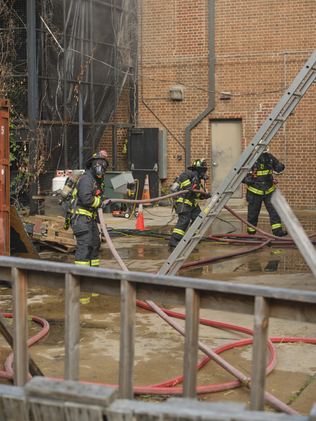 Feuerwehrleute in Helmen arbeiten daran, ein Gebäude Feuer zu löschen, umgeben von Ausrüstung wie Rohren, einem Metallzaun, einem Container und einem Verkehrskegel.