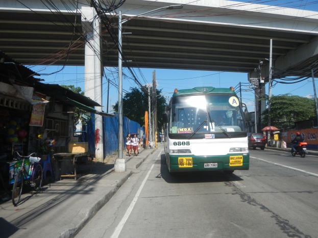 Ein grüner und weißer Bus fährt auf einer Straße neben einer Brücke, mit Strommasten und Drähten entlang der Straße, Fußgängern auf dem Gehweg, parkenden Fahrrädern, Geschäften, Bäumen und einem klaren Himmel im Hintergrund.