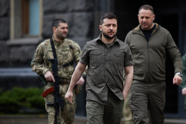 Ein Mann in militärischer Uniform, bei dem es sich mutmaßlich um den ehemaligen ukrainischen Präsidenten Viktor Janukowitsch handelt, wird von zwei bewaffneten Männern in militärischer Uniform vor einem Gebäude mit Pflanzen im Hintergrund eskortiert.