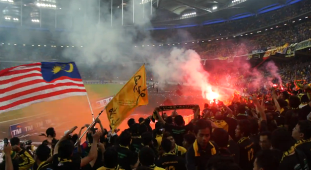 Eine große Menschenmenge in einem Stadion mit Bengalos, Fahnen und einer Kamera, mit Rauch und Schildern, auf denen "Malaysia vs Malaysia Live Streaming" steht.