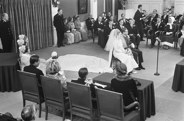 Schwarzes und weißes Foto einer Hochzeitszeremonie mit sitzenden und stehenden Menschen, einem Mann mit Mikrofon und einer Frau mit Blumenstrauß; ein Podium mit Mikrofon und Blumenschmuck ist auf beiden Seiten zu sehen.