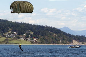 Eine Person, die über einem Gewässer gleitschirmfliegt, mit einem Boot auf der rechten Seite, Bäumen, Gebäuden, Hügeln und einem klaren blauen Himmel im Hintergrund.
