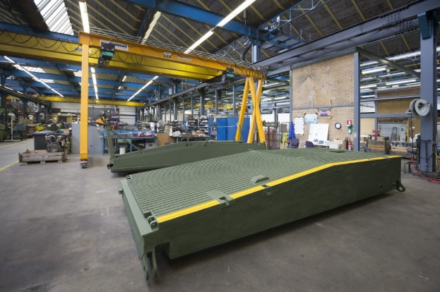 Großes Lagerhaus mit grünen Containern, Maschinen, Polen, Beleuchtung und Stangen; Hintergrund zeigt eine Wand mit Uhr und angehefteten Papieren.