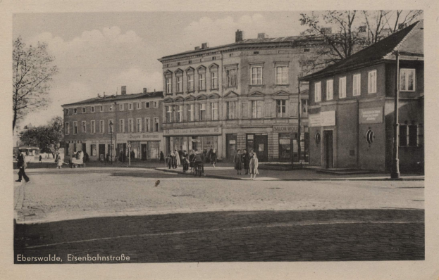 Altes Schwarz-Weiß-Foto einer Stadtstraße in Eberswalde, Eisenbahnstraße, mit Gebäuden, Bäumen, Menschen, Strommasten, Schildern und einem bewölkten Himmel, mit Text unten.
