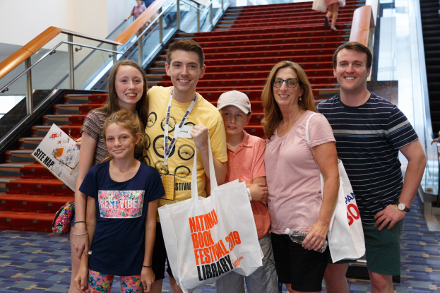 Eine Gruppe von Menschen, die vor einer Rolltreppe stehen, lächeln und Taschen halten, mit Treppen, Geländern und einer Wand im Hintergrund.
