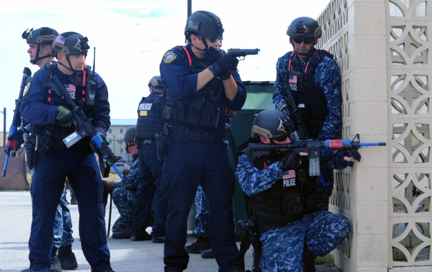 Gruppe von Polizisten in Helmen und Uniformen auf einer Brücke stehend, Waffen haltend, mit einer Wand zur Rechten und Gebäuden, Polen, Bäumen und Himmel im Hintergrund.