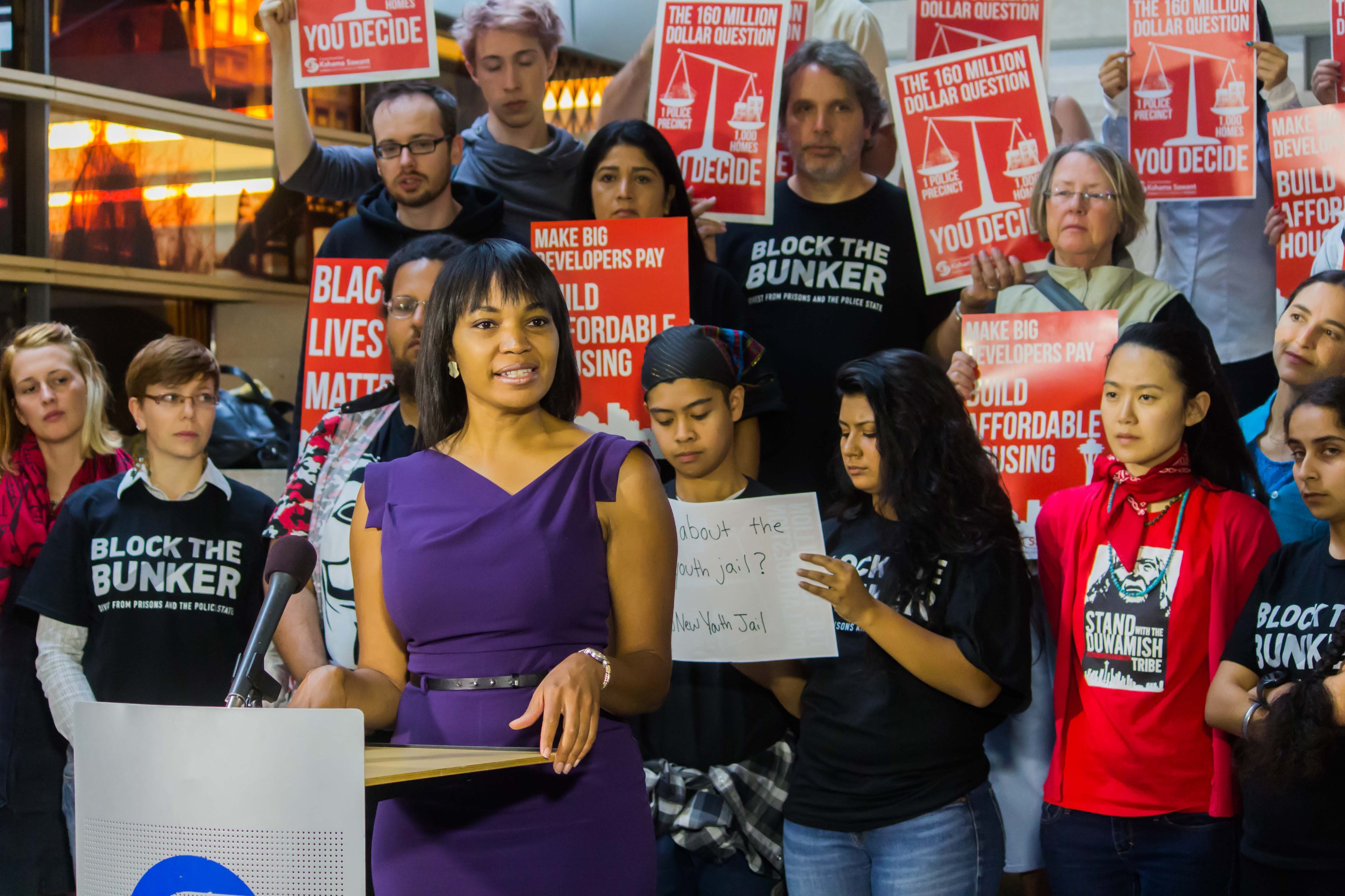 Eine Black Lives Matter-Aktivistin spricht vor einem Publikum mit Mikrofon, das Plakate hält, mit Lichtern und Gegenständen im Hintergrund.
