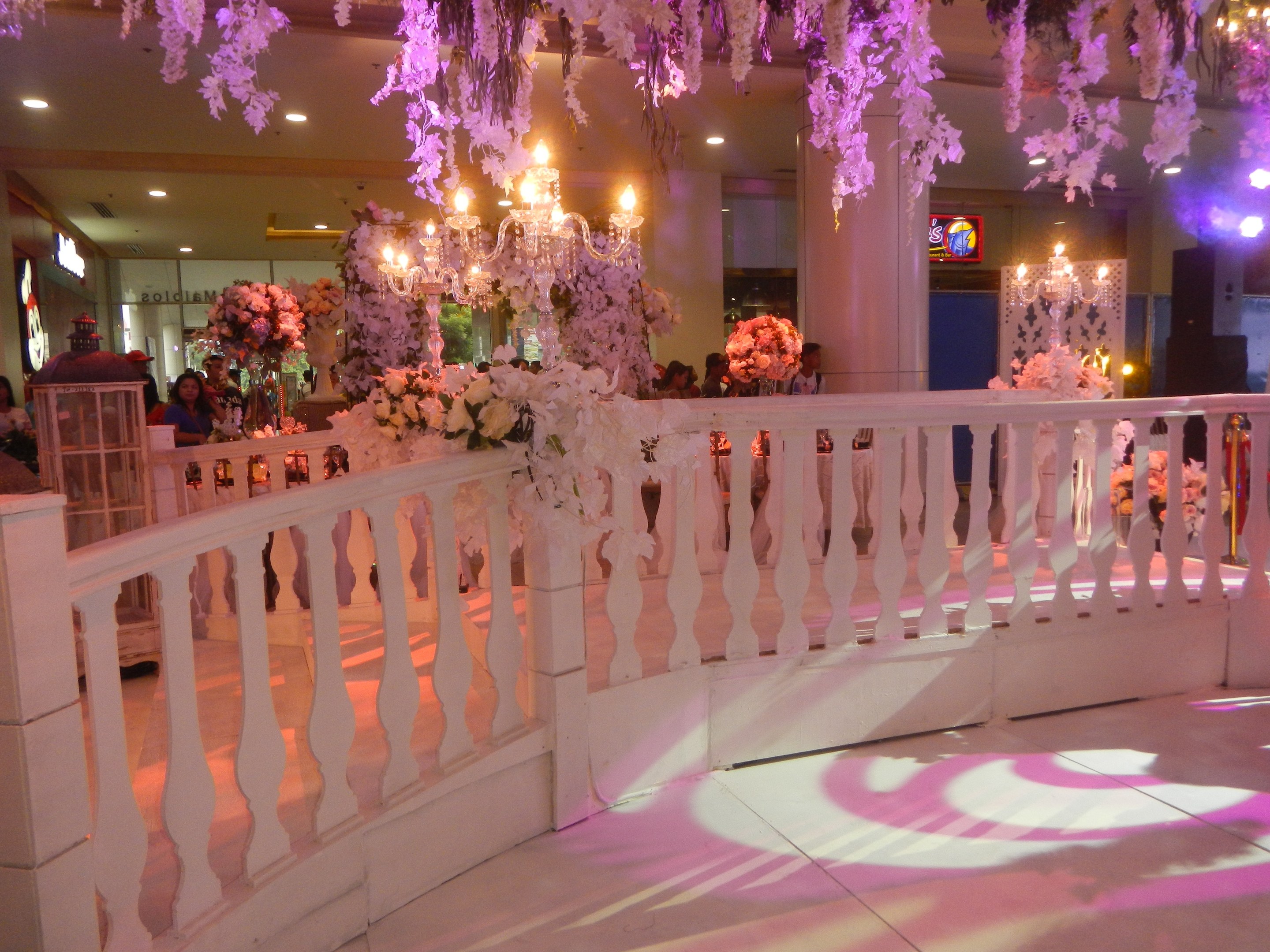 Weißes Geländer mit Blumen und Kronleuchtern geschmückt, umgeben von Menschen, Säulen und Lichtern im Hintergrund und ein Bildschirm auf der rechten Seite des Raums.